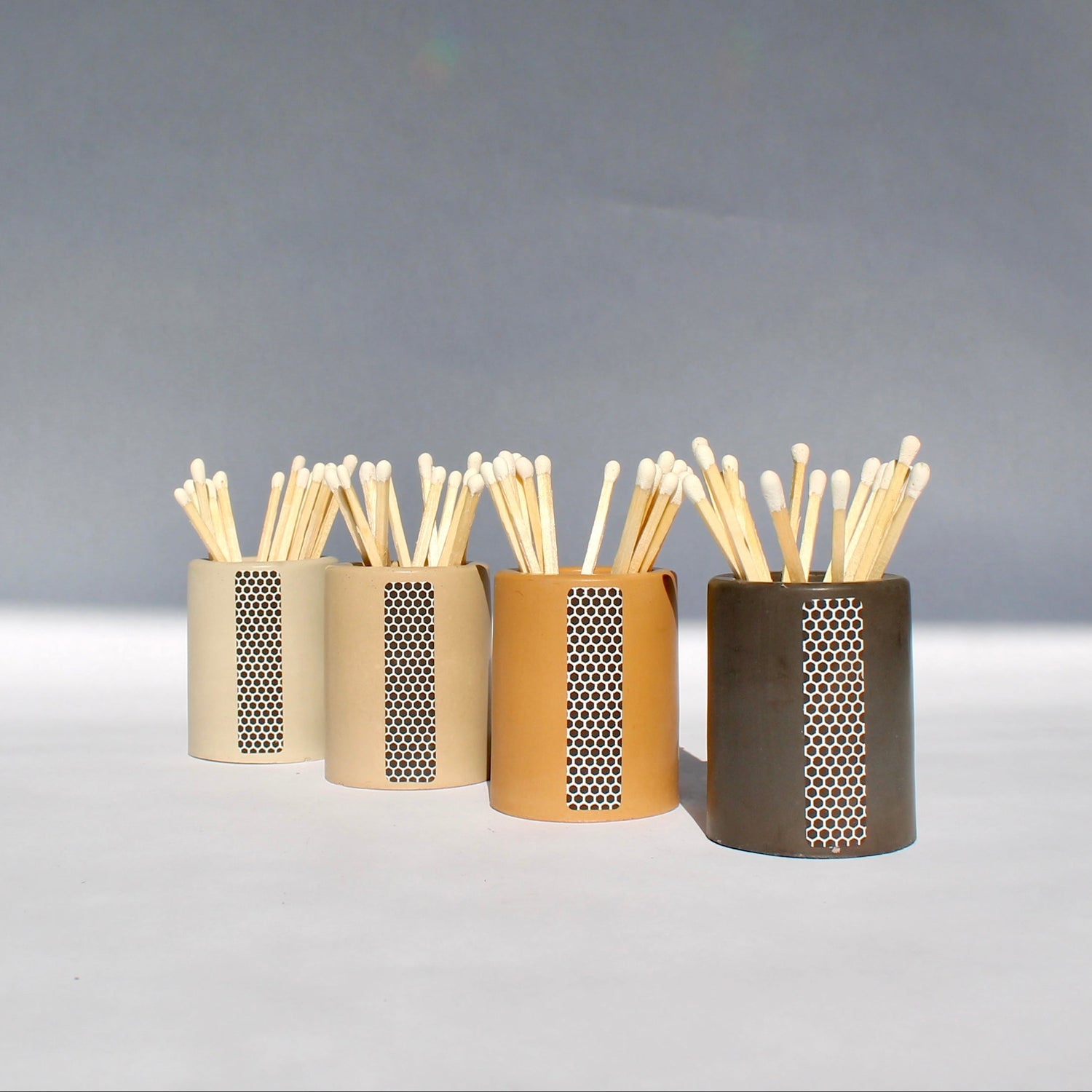 A collection of concrete match striker holders in beige, gray, and brown colors, filled with matches, displayed on a light background.