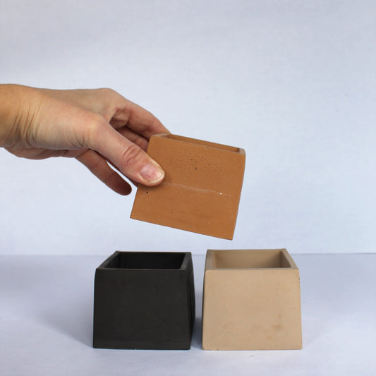 Hand holding a clay-colored square concrete planter above two other small square planters, one black and one beige, on a white background.