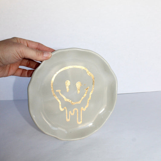 Hand holding a concrete decorative plate with a gold drippy smiley face design on a white background