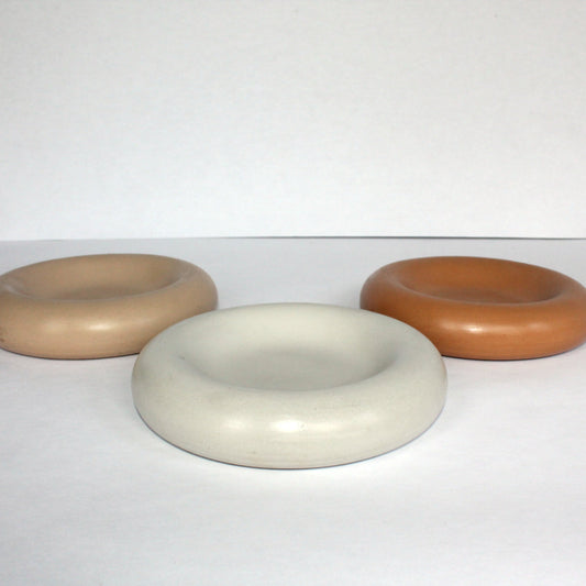 Three thick round concrete trays in beige, white, and terracotta on a white surface.