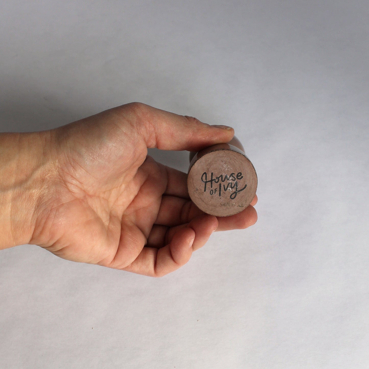 Hand holding concrete candle stick holder showing the bottom with 'House of Ivy' on a plain background