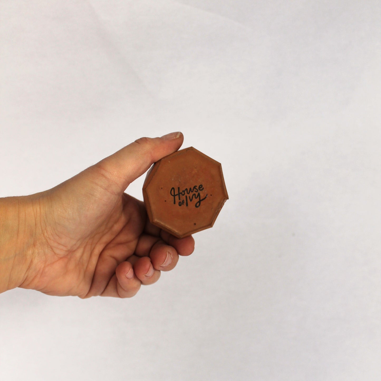 Hand holding a hexagonal terracotta object with 'House of Ivy' stamped on bottom against a white background.
