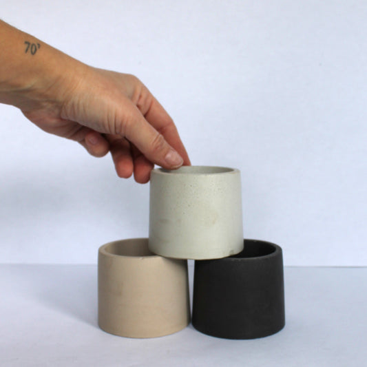Three concrete pots in beige, gray, and black being held by a hand on a white background