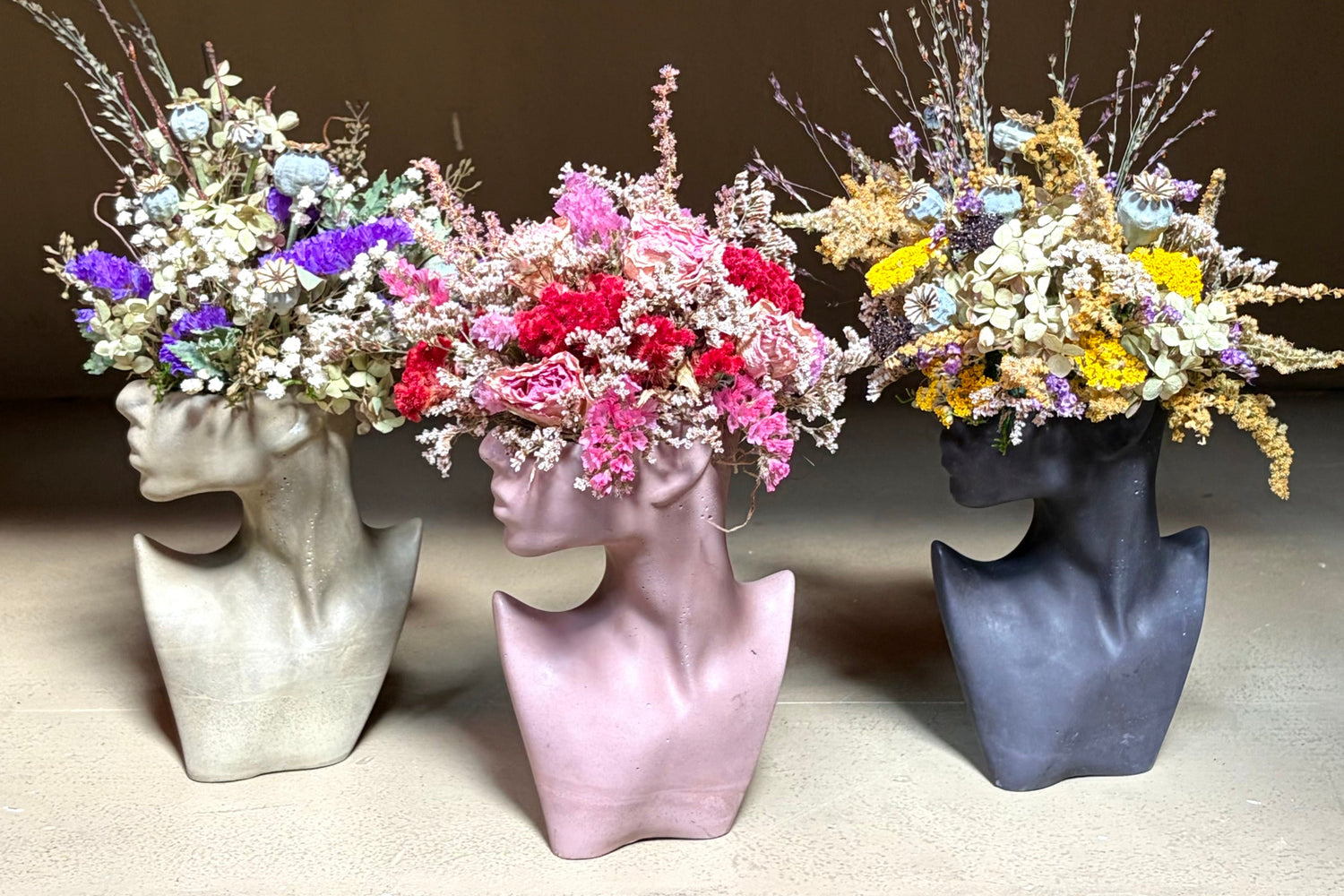 Three concrete female silhouette flower vases in sage green, mocha brown, and black, filled with dried flowers, arranged on a shelf against a brown background.