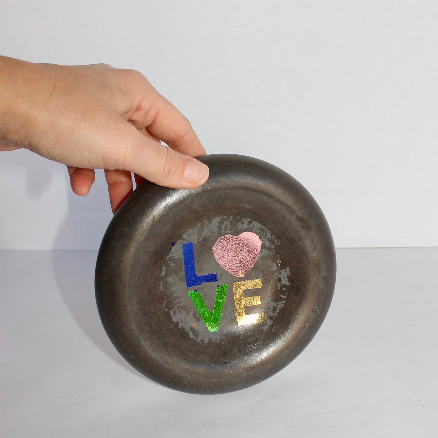 Hand holding a small thick round black concrete tray with colorful letters spelling 'LOVE' on a white background