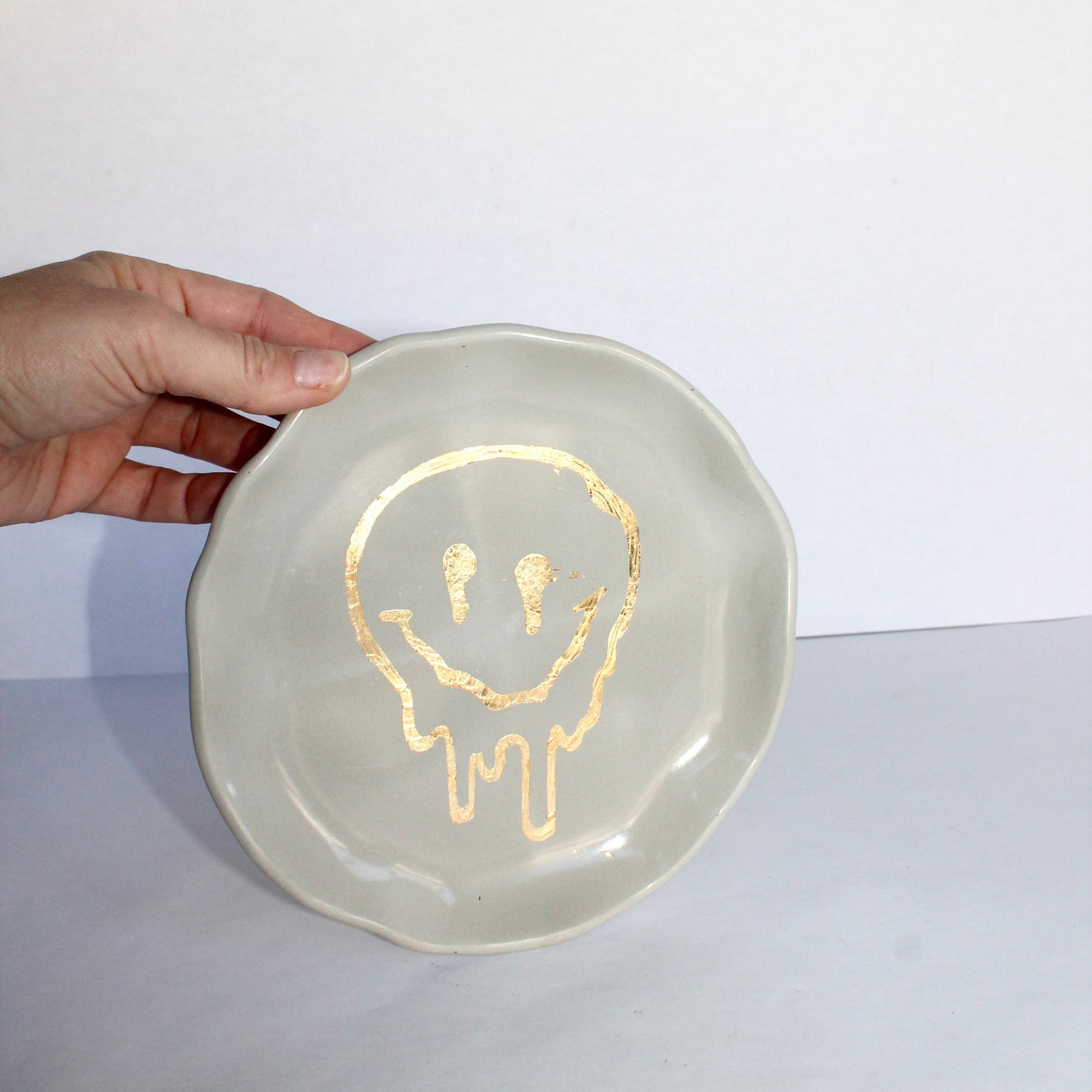 Hand holding a concrete decorative plate with a gold drippy smiley face design on a white background