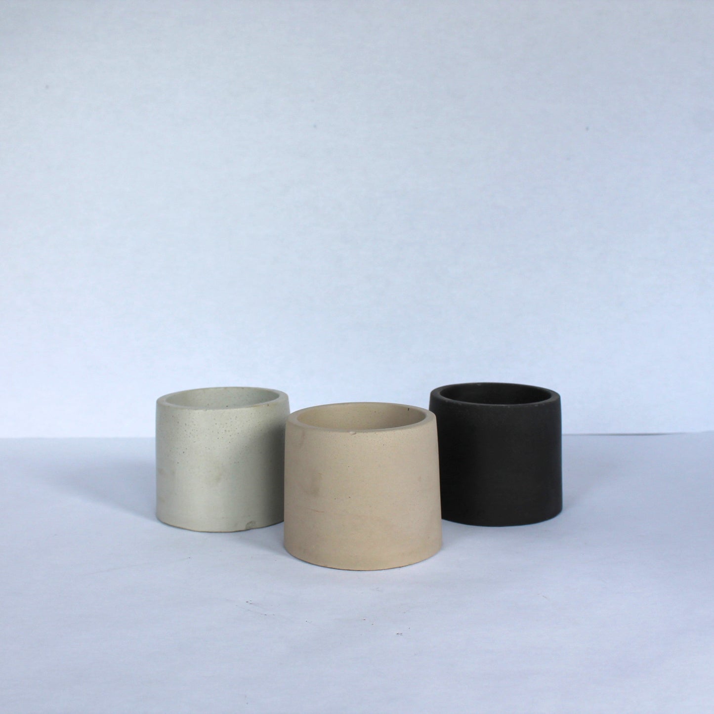 Three cylindrical concrete pots in beige, gray, and black on a white background