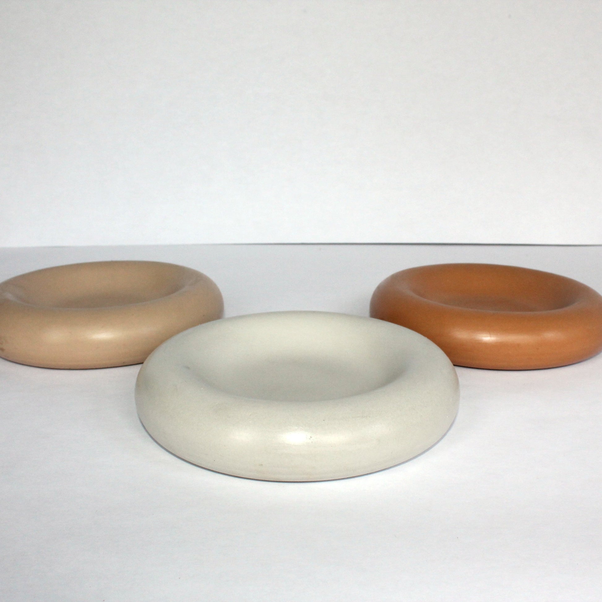 Three thick round concrete trays in beige, white, and terracotta on a white surface.