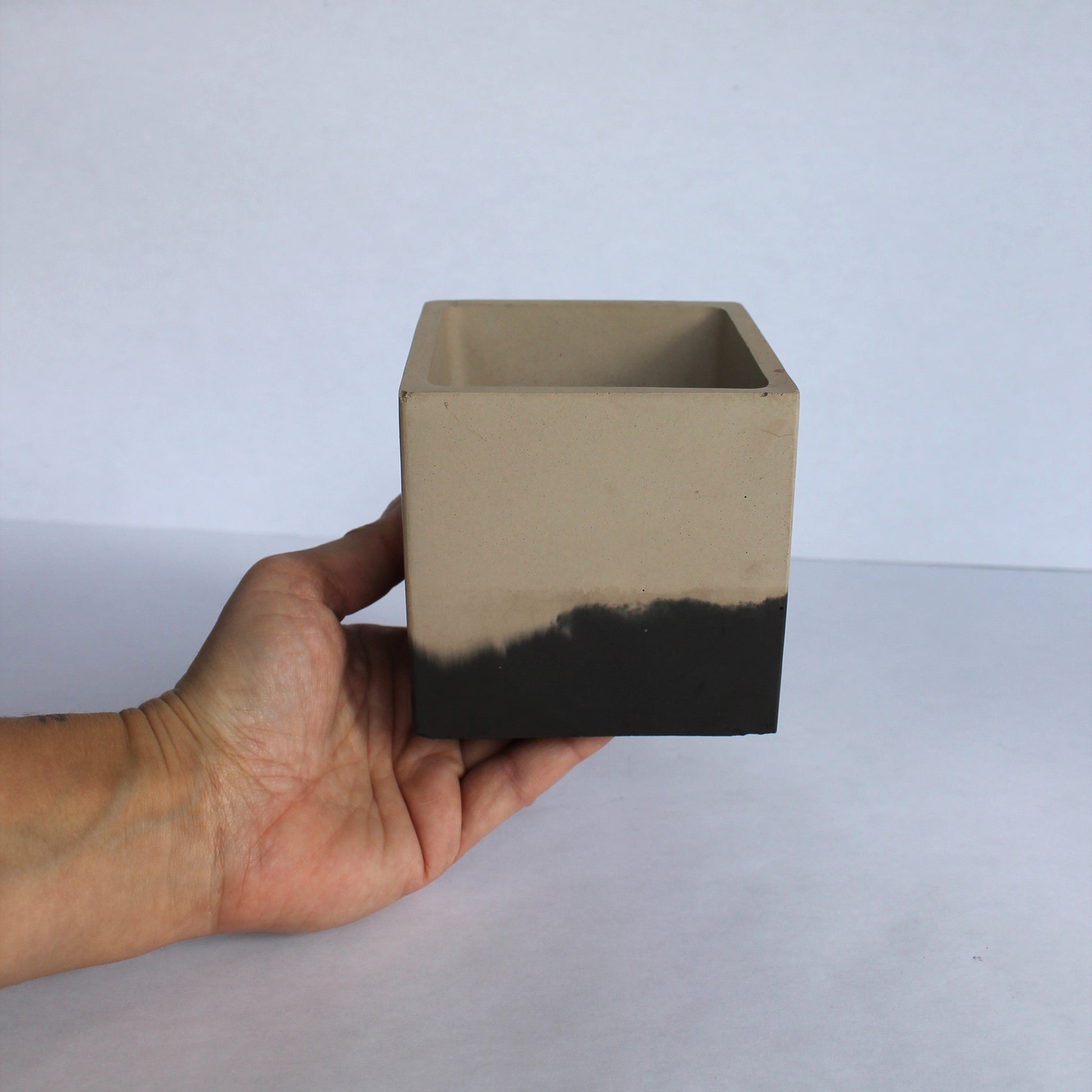 Hand holding a square concrete planter with black base against a white background